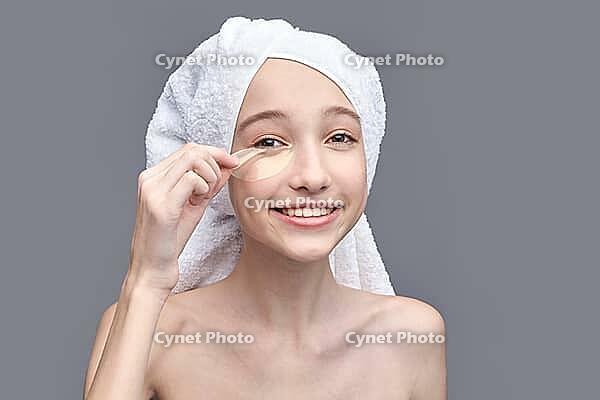 Young caucasian female applying under eye patch with towel on head against gray background [IBR123629771]
