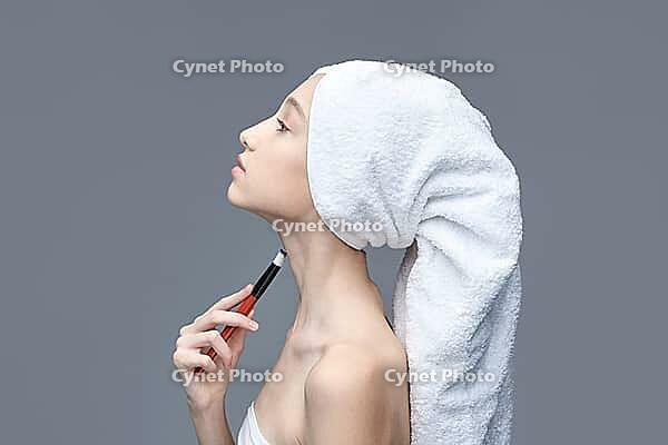 Young caucasian female applying makeup with brush on neck wrapped in towel against gray background [IBR123629773]