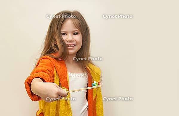 Caucasian young girl in orange bathrobe holding toothbrush with multicolored toothpaste [IBR123629774]
