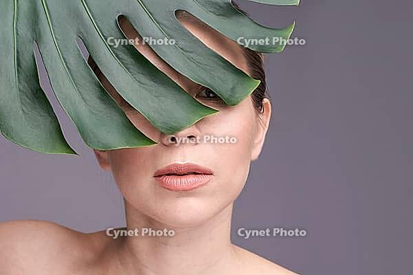 Caucasian female with monstera leaf partially covering face, close up portrait on gray background [IBR123629776]