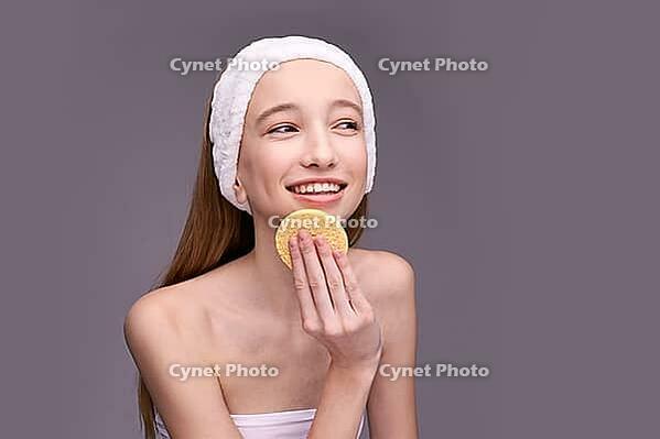 Smiling caucasian female teen using facial sponge for skin care routine with headband on grey background [IBR123629777]