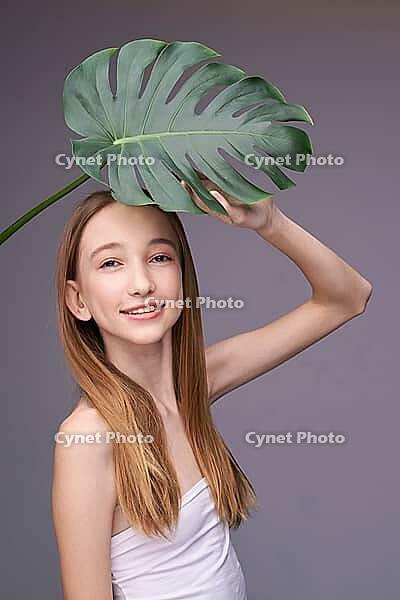 Young caucasian female holding monstera leaf in studio on gray background with long blonde hair and white top [IBR123629778]