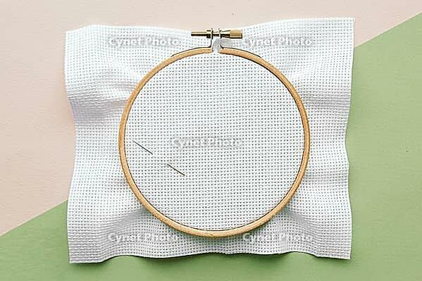Wooden embroidery hoop with empty white canvas cloth and needle on pastel background with light pink and light green colors, ready for cross-stitching [IBR123638370]