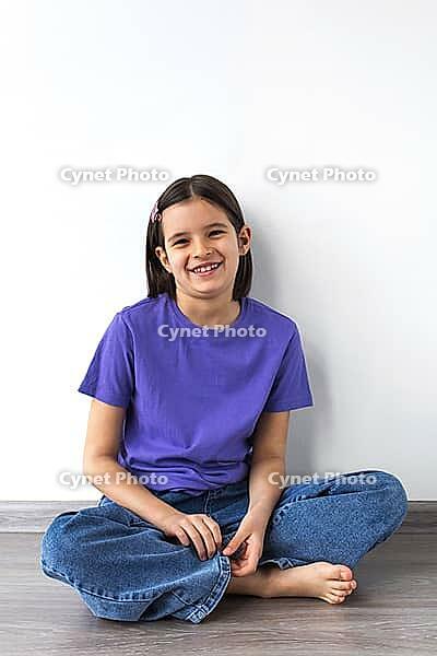 Smiling white brunette girl aged 7-8 in purple t-shirt and jeans sitting barefoot on wooden floor against white wall [IBR123638371]