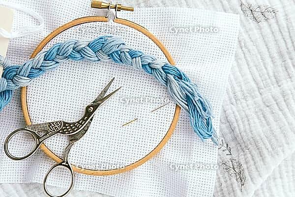 Embroidery hoop with white canvas, braid of blue thread, stork-shaped scissors and needles on soft textile background, representing craftsmanship, patience and creativity, cross stitch concept [IBR123638372]