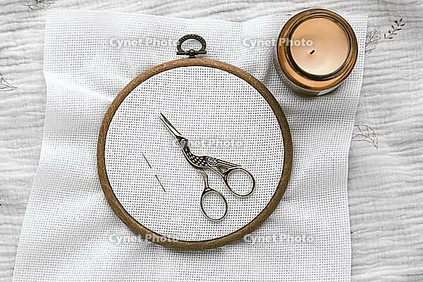 Embroidery hoop with white canvas, vintage stork-shaped scissors and needle on textured fabric, next to a lit candle in warm cozy atmosphere, representing creativity and handmade crafts, cross stitch [IBR123638373]
