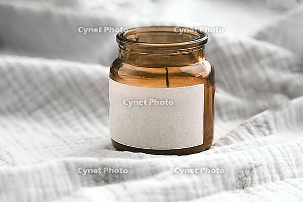 Brown glass candle jar with blank label standing on soft crumpled fabric in natural light, minimalistic cozy composition for branding or product display [IBR123638375]