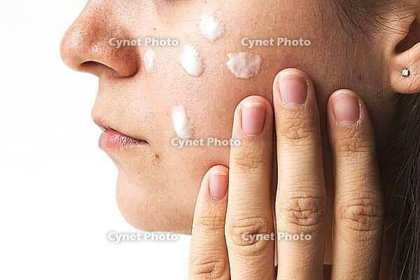 Close-up of young woman applying skincare cream to her cheek with fingers, visible texture of skin and cream on white background, concept beauty, skincare, self-care [IBR123638376]