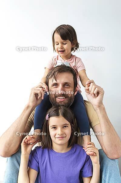 Father with two daughters posing together, smiling having fun. younger girl sits on his shoulders, while older one holds his hands. Happy moment in bright interior, concept of fatherhood, vertical [IBR123638377]