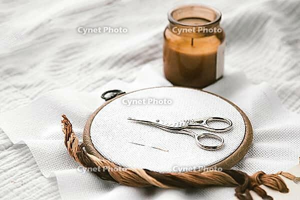 Embroidery hoop with white fabric, vintage scissors, and a needle lying on top, accompanied by a burning candle in a cozy setting. Aesthetic composition of handmade craft and relaxation [IBR123638379]
