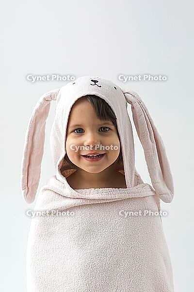 Smiling child wrapped in soft pink towel with bunny ears, looking directly at camera against a white background. hood has an embroidered rabbit face, creating a cute and cozy appearance, vertical [IBR123638381]