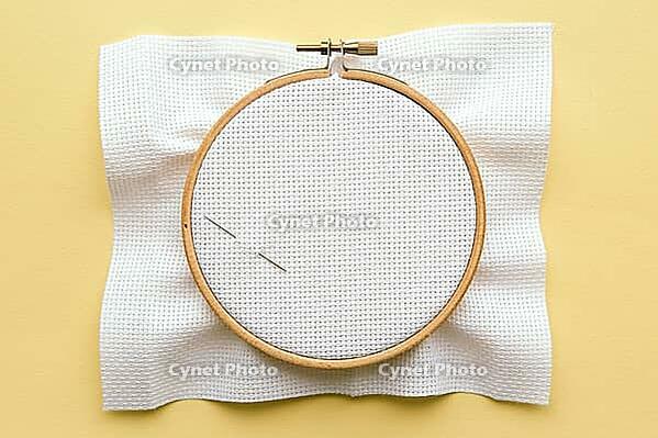 Wooden embroidery hoop with white fabric and two needles on a yellow background, prepared for cross-stitching and needlework concept [IBR123638382]
