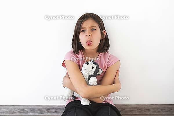 Child girl 7-8 years in pink t-shirt sits against white wall, hugging plush toy and sticking out her tongue with stubborn expression. She appears playful and defiant. Concept of childhood emotions [IBR123638383]