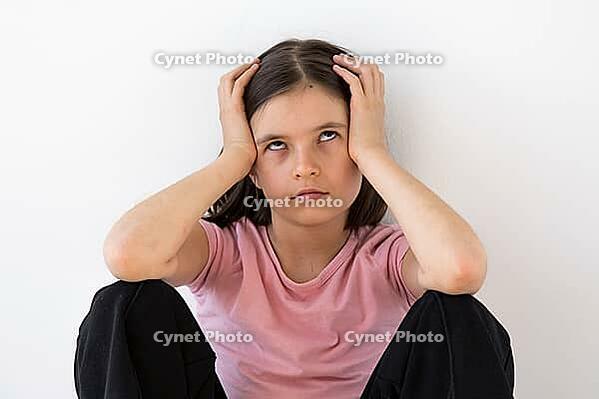 A young girl 7-8 year in a pink shirt sits against a white wall, rolling her eyes and holding her head in frustration, symbolizing boredom, annoyance, or stress [IBR123638387]