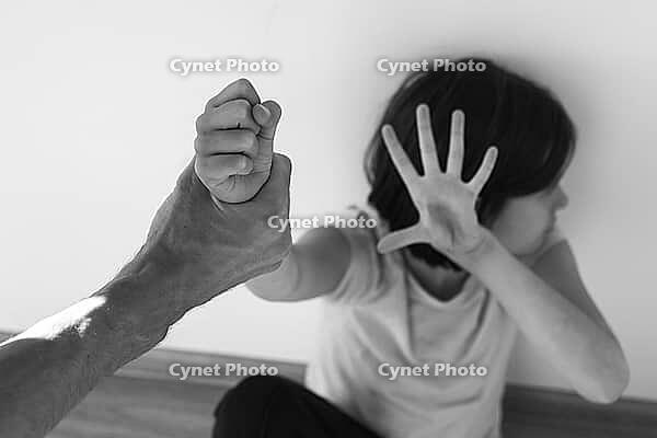 Black and white photography frightened young girl in a pink shirt sits against a wall, raising her hand in defense while an adult tightly grips her wrist, symbolizing domestic violence and child abuse [IBR123638388]