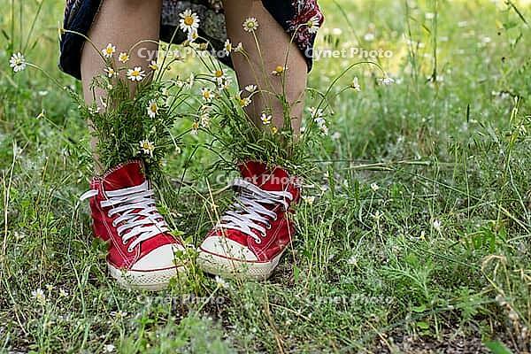 Upturned skirt and naked legs in red shoes with a bouquet of white camomiles in green grass. Summer concept [IBR123639491]