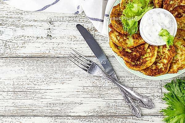 Fritters from zucchini with sour cream sauce and greens. Vegetable pancakes of courgettes with green salad on white wooden background. Top view with copy space [IBR123639579]