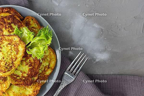Fritters from zucchini in grey plate with sour cream sauce and greens. Vegetable pancakes of courgettes with green salad on grey background. Top view with copy space [IBR123639583]
