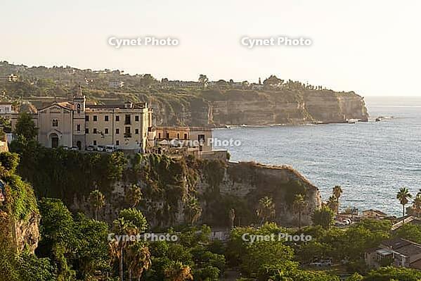 Tropea, Italy - 27.07.2023: Picturesque view of historic building perched on cliff, overlooking ocean and surrounded by lush greenery with distant cliffs and coastline in background [IBR123646525]