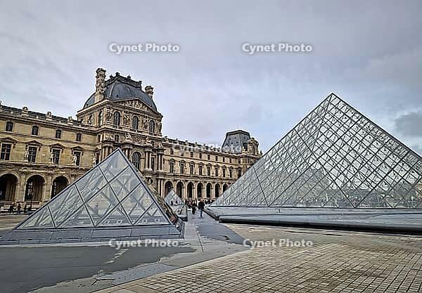 Paris, France - December 10, 2025: The ornate Renaissance facade and the iconic glass Pyramid of the Louvre Museum with multiple tourists scattered across the vast cobblestone courtyard [IBR123895592]