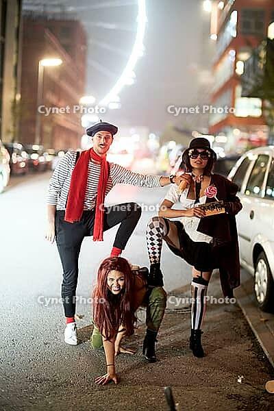 Dominance hierarchy of three friend in costume posing playfully under city lights on an urban street at night, nightlife, dog-like attitude of submissive playful woman crawling on street floor while her male friend steps on her back with his foot like her [IBR123999807]