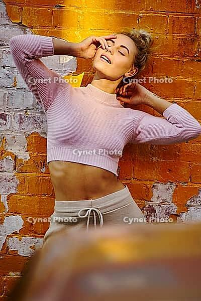 Sexy attraction, a smiling young woman poses playfully against a bright orange brick wall under sunlight, feeling sexy, desires, sexiness, attraction, stretching, stretch, insightful, attractive, wistful, leisure, casual, informal, relaxed, laid-back, ten [IBR124192783]