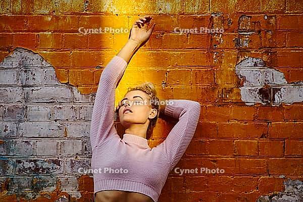 Laid-back young woman joyfully stretches against a sunlit brick wall in a pink sweater, by sunshine enlightened face, sunning, Vitamin D, summer, warmth, facial, face, skin, health, resting, restful, pause, relax, relaxation, relaxing, wistufl, longing, d [IBR124192784]