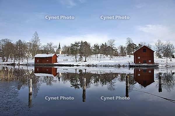 Classic red Swedish wooden house on the water with the reflection of the Borgvik church tower. Winter scene with snow and traditional Falunred architecture in Värmland, Sweden [IBR124200516]