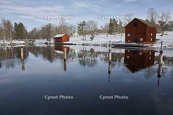 Classic red Swedish wooden house on the water with the reflection of the Borgvik church tower. Winter scene with snow and traditional Falunred architecture in Värmland, Sweden [IBR124200550]