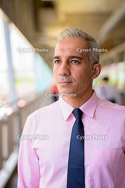 Portrait of handsome Persian businessman with gray hair at the footbridge in the city [IBR124309083]