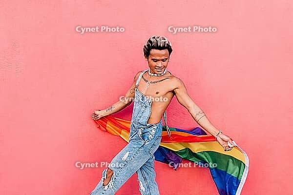 Young guy in denim overalls having fun holding rainbow LGBT flag [IBR124491532]