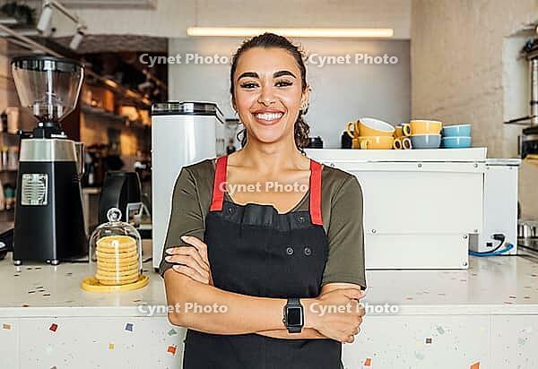 Female barista standing arms crossed in a cafe. Smiling waitress looking at camera while standing in front of a counter [IBR124491549]