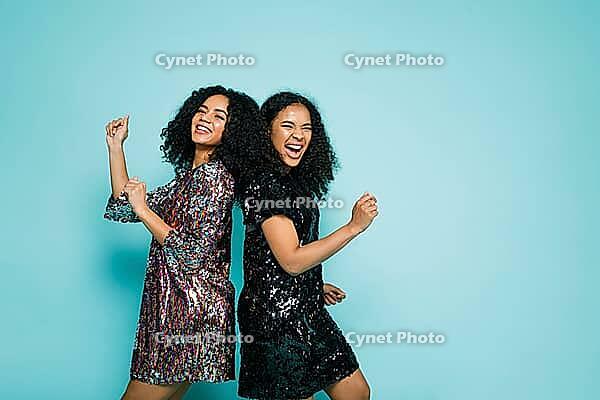 Female friends dancing back to back against blue background, wearing stylish clothes [IBR124491555]