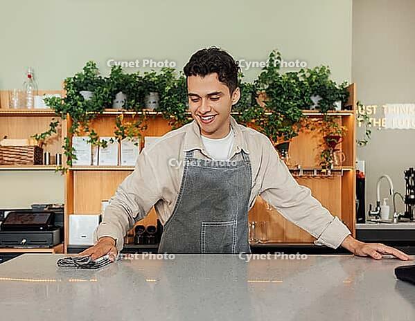 Smiling barista cleaning a counter in a cafe with a towel. Young male in an apron working as a bartender [IBR124491558]
