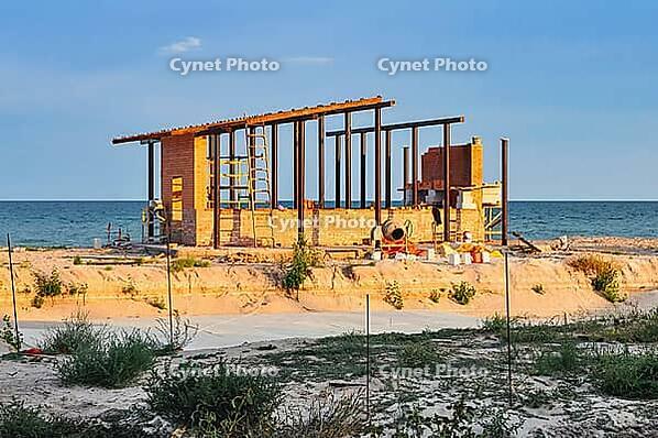 A brick building is under construction on the sandy beach near the sea, at sunset. Construction equipment can be seen [IBR124491563]