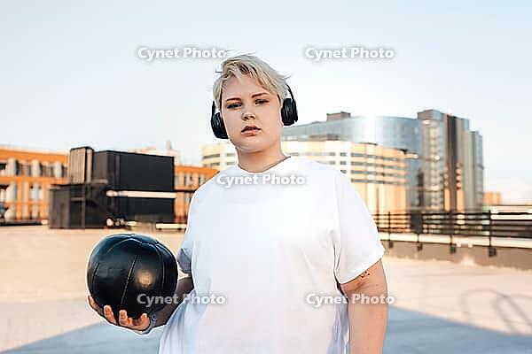Confident female standing on the roof with medicine ball and headphones on her head [IBR124491575]