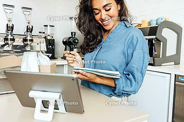 Smiling woman holding a note book in coffee shop. Cafe owner standing at table [IBR124491581]