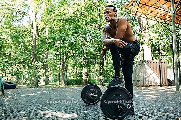 Bare-chest bodybuilder resting after deadlifting exercises at the sports ground. Young athlete stands with one leg on the barbell [IBR124491599]