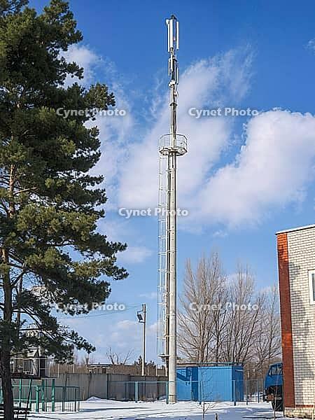 A tall cell tower with antennas rises against a bright blue sky dotted with clouds in a winter landscape [IBR124491601]