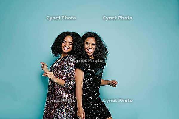 Female friends dancing back to back in studio against blue background [IBR124491627]