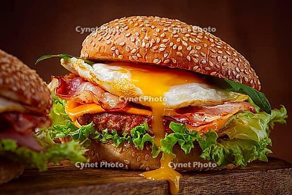 Two juicy burgers sit on a wooden board, each filled with lettuce, bacon, cheese, and fried eggs [IBR124552175]