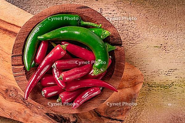 Bright green and red peppers are neatly placed in a round wooden bowl on a wooden board. The board has a natural finish, showing the grain of the wood [IBR124552189]