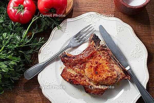 A pork chop rests on a white plate with a fork and knife. Fresh tomatoes and green herbs surround the plate on a wooden table [IBR124552205]