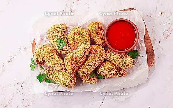 A wooden board holds several golden chicken drumsticks arranged neatly. A small bowl of red dipping sauce is nearby. Fresh parsley adds a touch of color [IBR124552219]