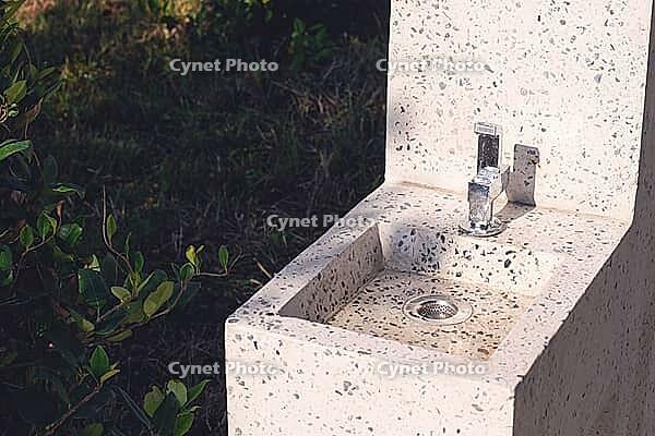 This outdoor sink features a sleek terrazzo design, with a shiny faucet and a simple drain. It is situated in a landscaped garden, bathed in warm sunlight [IBR124552247]