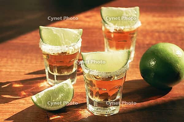 Three glasses with tequila and salt on the rims are arranged on a wooden table. Each glass has a slice of lime on top. A lime sits in the background under natural light [IBR124552248]