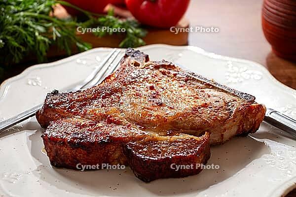 A pork chop rests on a white plate with a fork and knife. Fresh tomatoes and green herbs surround the plate on a wooden table [IBR124552264]