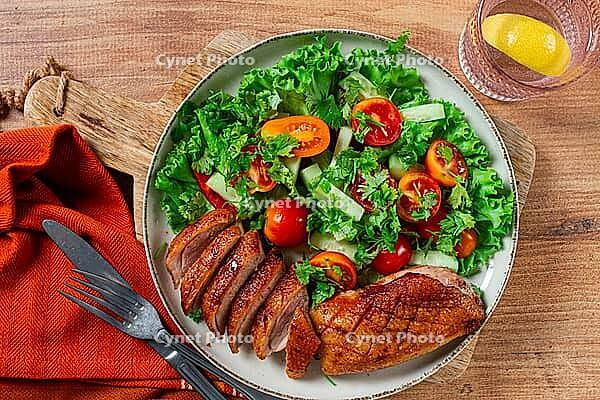 Delicious grilled duck breast served with fresh garden salad and cherry tomatoes at a rustic dining table [IBR124585337]