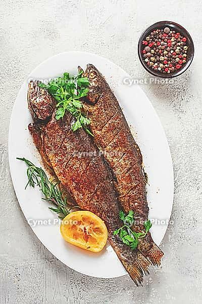 Two grilled fish are placed on a white dish. Fresh lemon slices and herbs are arranged beside them [IBR124585341]