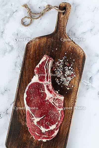 Raw rib eye steak, cowboy steak, wooden chopping board, marble table, top view, no people [IBR124585354]